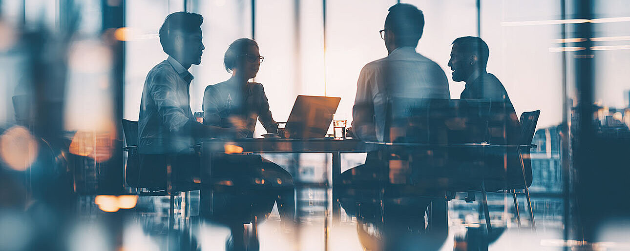 © hao - AdobeStock.com Vier Personen sitzen in einem modernen Konferenzraum mit großen Fenstern und besprechen sich am Tisch.