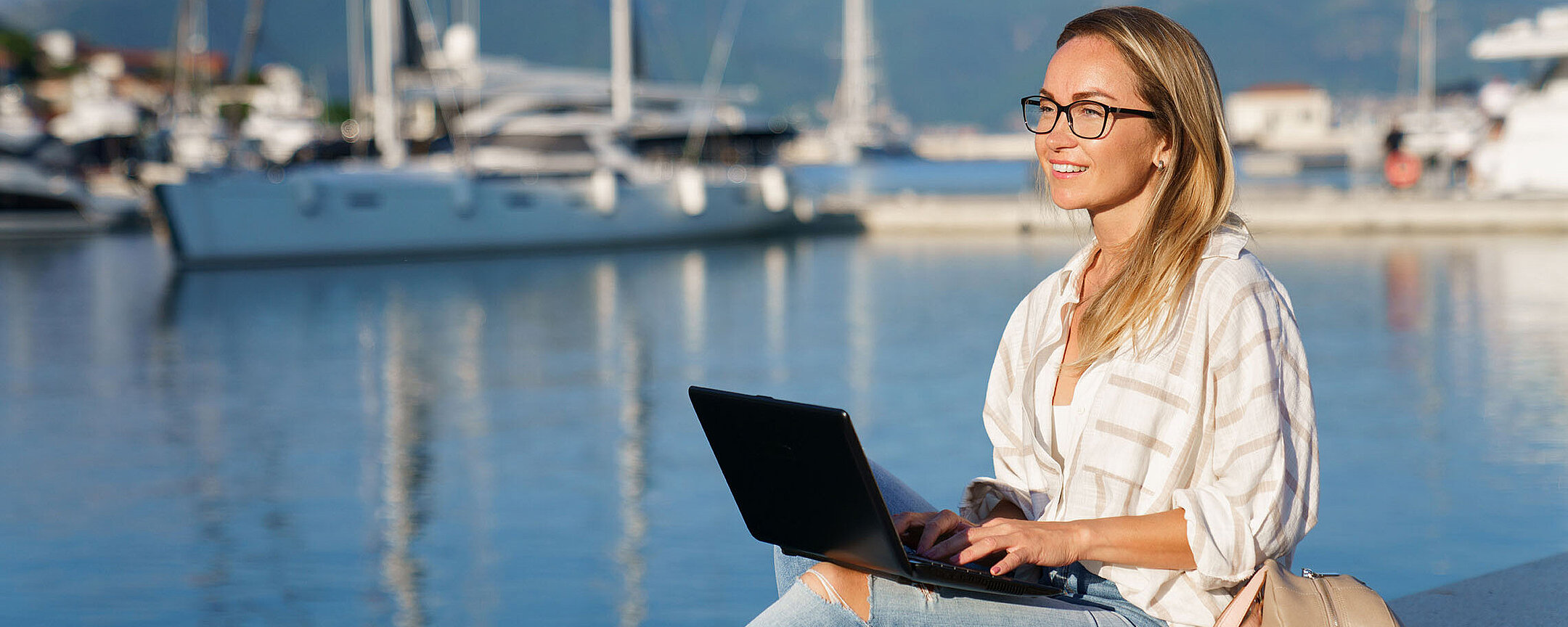 © Mary Anlens - AdobeStock.com Frau arbeitet mit Laptop am Hafen vor Segelbooten und ruhigem Wasser bei Sonnenschein.