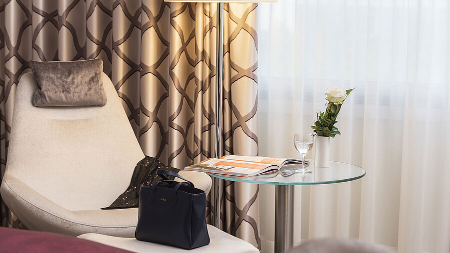 Zimmerdetail Detail eines Classic Zimmers im Maritim Hotel Bonn mit gemütlichem Sessel, Vorhang, Tisch, Glas, Rose und einer Tasche.
