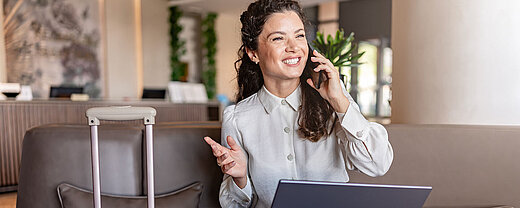 © lordn - AdobeStock.com Geschäftsfrau sitzt mit Laptop auf dem Sofa in einer Hotel-Lobby, telefoniert lächelnd und hat einen Koffer neben sich