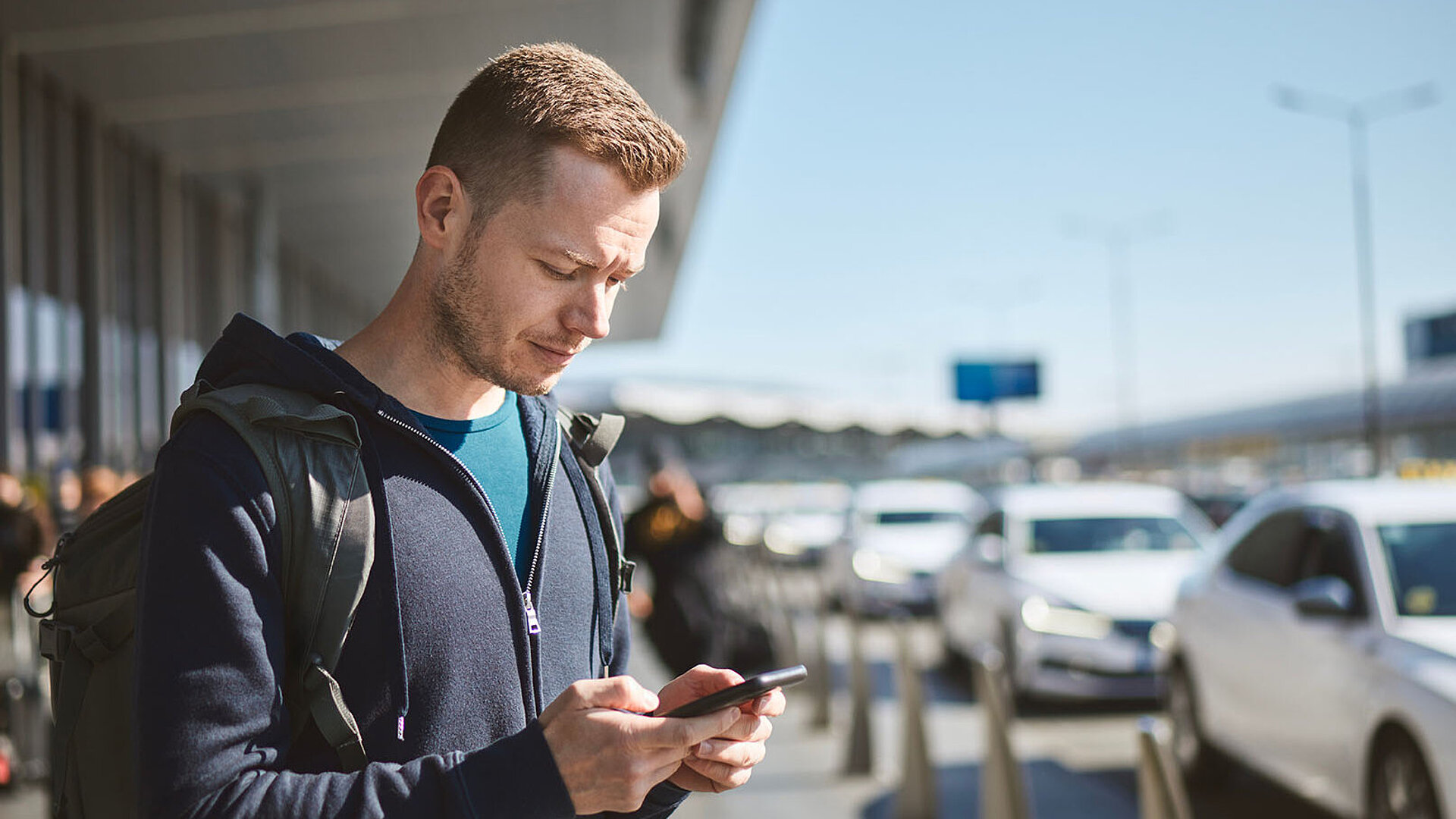 © Chalabala - AdobeStock.com Mann mit Rucksack nutzt Smartphone vor Flughafen, bereit für Transfer oder Taxi