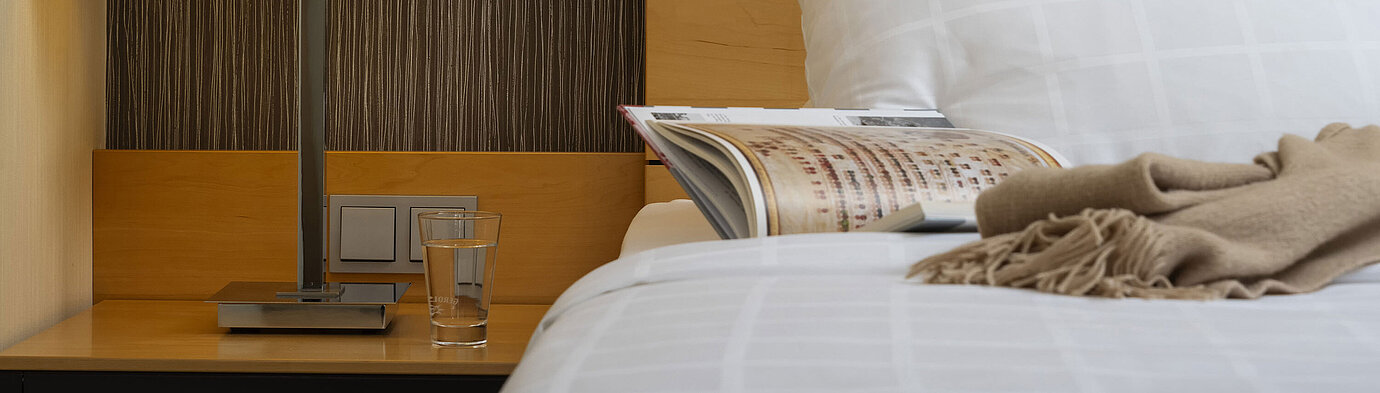 Classic Zimmer Detailaufnahme eines Nachttisches im Maritim Hotel Düsseldorf mit Lampe, Glas Wasser und Buch auf einem Bett.