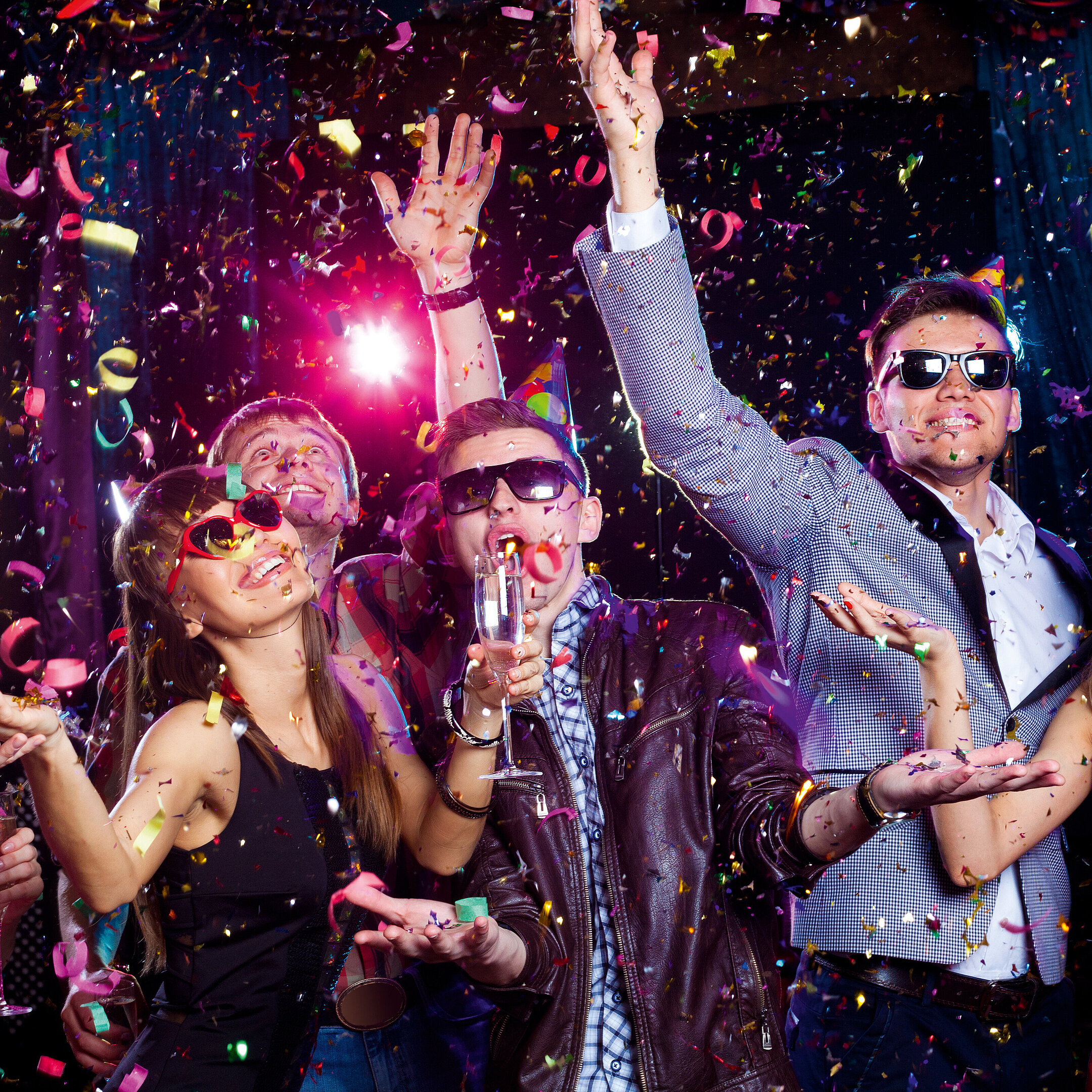 © yanlev - AdobeStock.com Fröhliche junge Leute feiern mit Partyhüten, Sonnenbrillen, Sektgläsern und Konfetti in bunter Clubatmosphäre