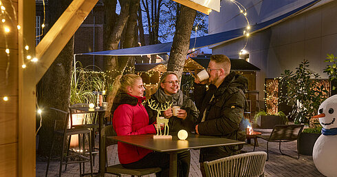 Maritim Weihnachtsdorf Drei Personen genießen heiße Getränke auf dem festlich geschmückten Weihnachtsmarkt im Maritim Darmstadt mit Lichterketten.