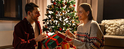 © Syda Productions - AdobeStock.com Zwei Personen sitzen vor einem geschmückten Weihnachtsbaum und tauschen liebevoll verpackte Geschenke aus.