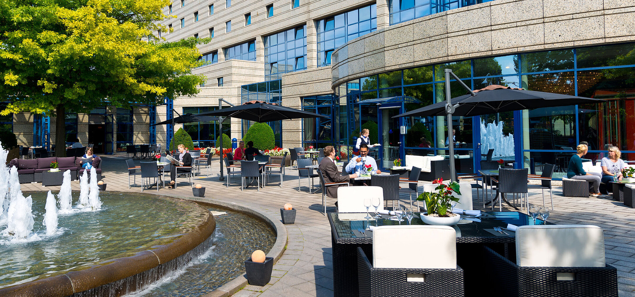 Terrasse Terrasse des Maritim Hotels Bonn mit Sitzbereichen, Springbrunnen und Gästen, die im Freien speisen und entspannen.