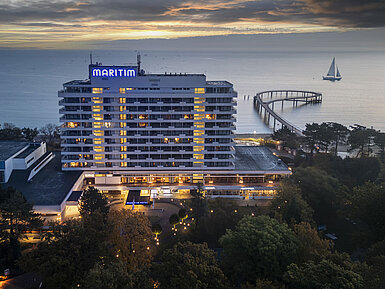 Außenansicht Abendaufnahme des Maritim Seehotels mit beleuchteter Seebrücke und einem Segelboot im Hintergrund bei Sonnenuntergang.
