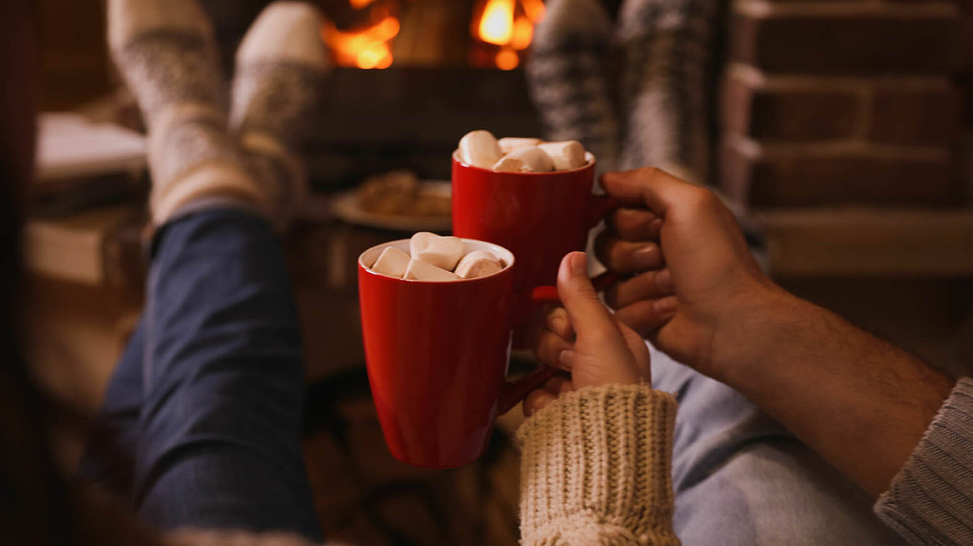 © New Africa - AdobeStock.com Zwei Personen entspannen am Kamin und halten rote Tassen mit Marshmallows in einer gemütlichen Winteratmosphäre.