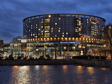 Außenansicht bei Nacht Maritim Hotel Frankfurt bei Nacht mit beleuchteter Glasfassade und Spiegelung im ruhigen Teich davor.