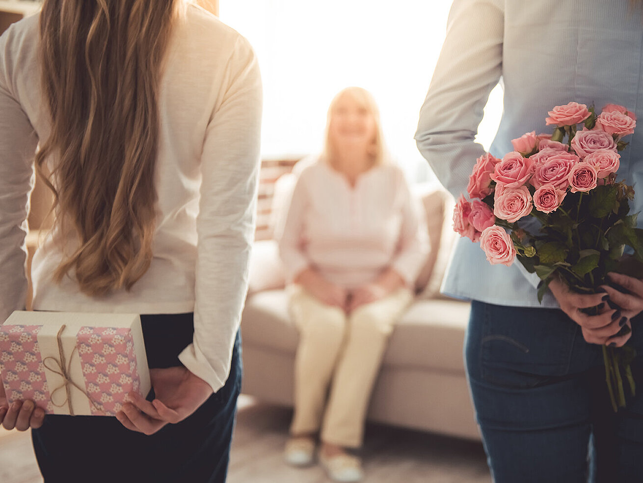 Muttertag © VadimGuzhva - AdobeStock.com Zwei Töchter überraschen ihre Mutter mit Blumen und Geschenk im Wohnzimmer