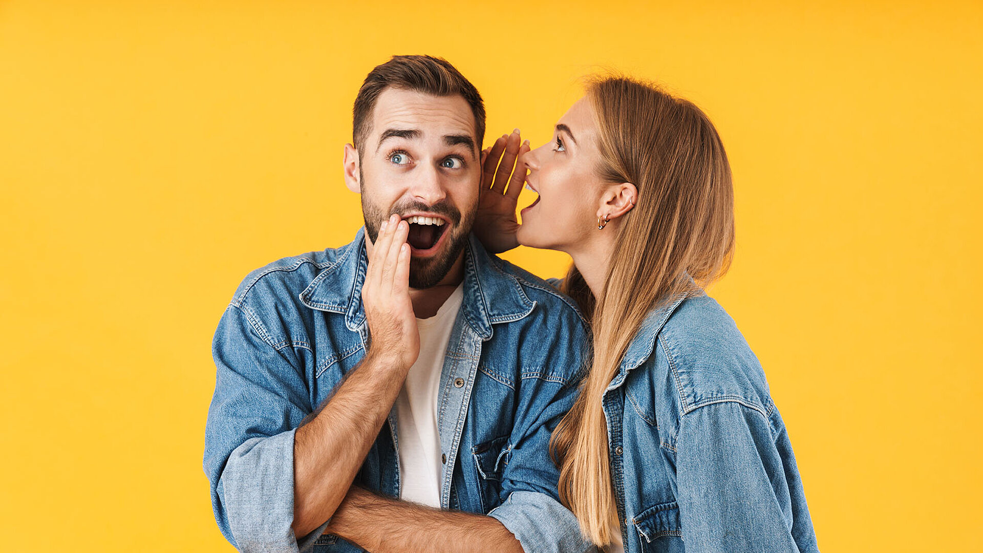 © Drobot Dean - AdobeStock.com Frau flüstert Mann ein Geheimnis ins Ohr, beide lachen und tragen Jeansjacken vor gelbem Hintergrund