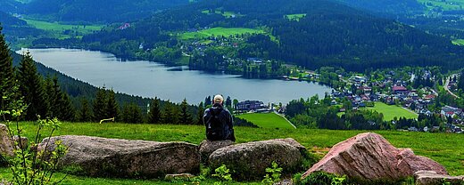 Schwarzwald Wanderer mit Rucksack sitzt auf Felsen und schaut auf den Titisee und das Maritim Hotel im Schwarzwald