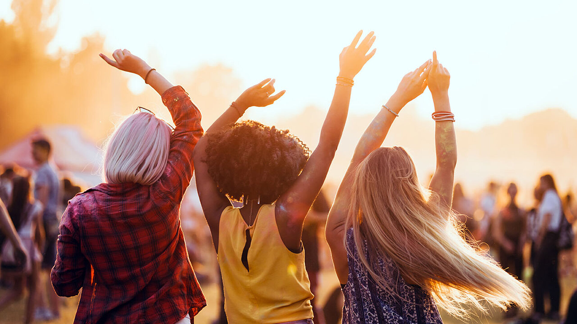© leszekglasner - AdobeStock.com Drei Freunde tanzen mit erhobenen Armen im Sonnenuntergang bei einem Musikfestival im Freien.