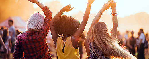 © leszekglasner - AdobeStock.com Drei Freunde tanzen mit erhobenen Armen im Sonnenuntergang bei einem Musikfestival im Freien.