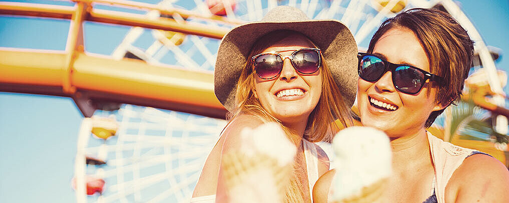 © oneinchpunch - AdobeStock.com Zwei Frauen essen lachend ein Eis. Im Hintergrund ist ein Riesenrad auf einem Jahrmarkt bei Sonnenschein zu sehen