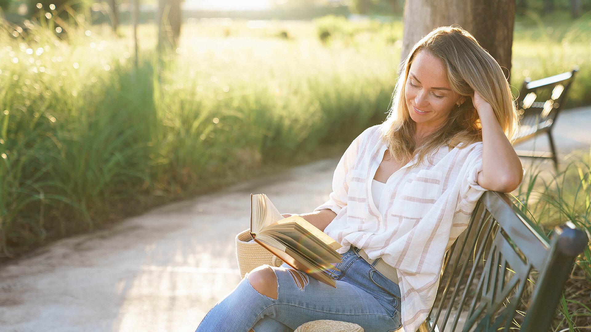 © Marina April - AdobeStock.com.jpg Frau liest entspannt ein Buch auf einer Parkbank im Sonnenschein – Auszeit in der Natur genießen