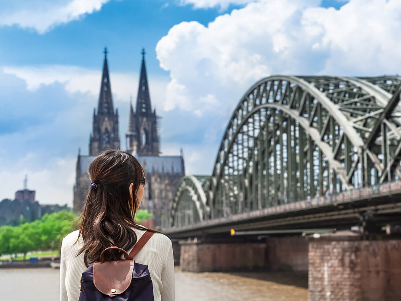 © 75tiks - AdobeStock.com Frau mit Rucksack blickt auf den Kölner Dom und die Hohenzollernbrücke am Rhein
