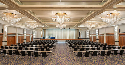 Fürst-von-Metternich-Saal Fürst-von-Metternich-Saal mit großer Bühne, Reihenbestuhlung, eleganten Kronleuchtern und hellem Ambiente.