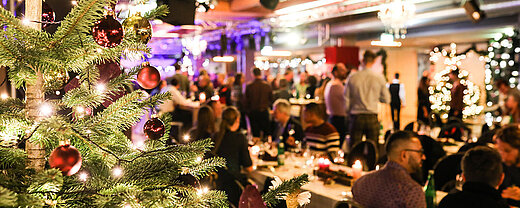 © bilderstöckchen - AdobeStock.com Weihnachtsfeier mit festlich geschmücktem Baum und Gästen im festlichen Saal