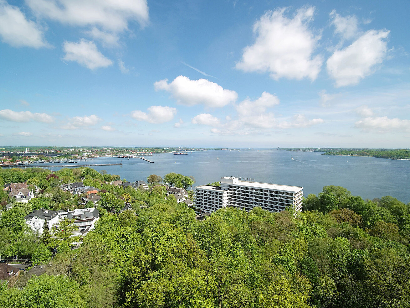 Außenansicht Luftaufnahme des Maritim Hotels Kiel, umgeben von grüner Landschaft und der Kieler Förde, bei strahlend blauem Himmel.
