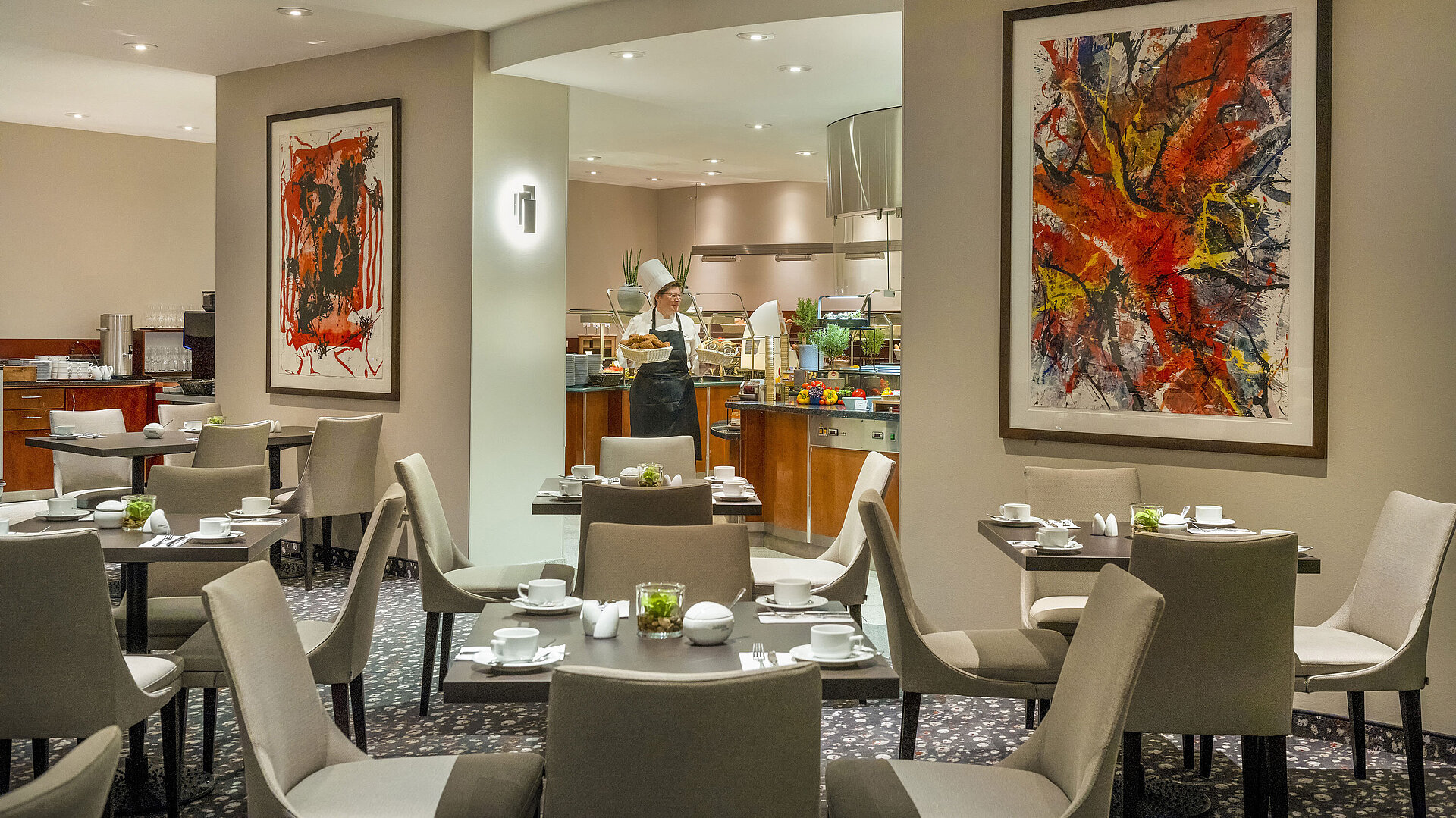 Restaurant Galerie Restaurant im Maritim proArte Hotel Berlin mit moderner Kunst an den Wänden, gedeckten Tischen und einem Koch am Buffet.