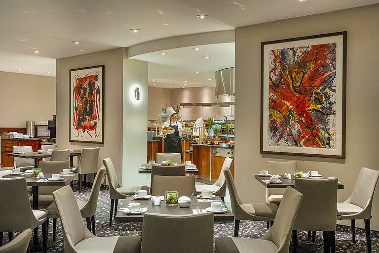 Restaurant Galerie Restaurant im Maritim proArte Hotel Berlin mit moderner Kunst an den Wänden, gedeckten Tischen und einem Koch am Buffet.