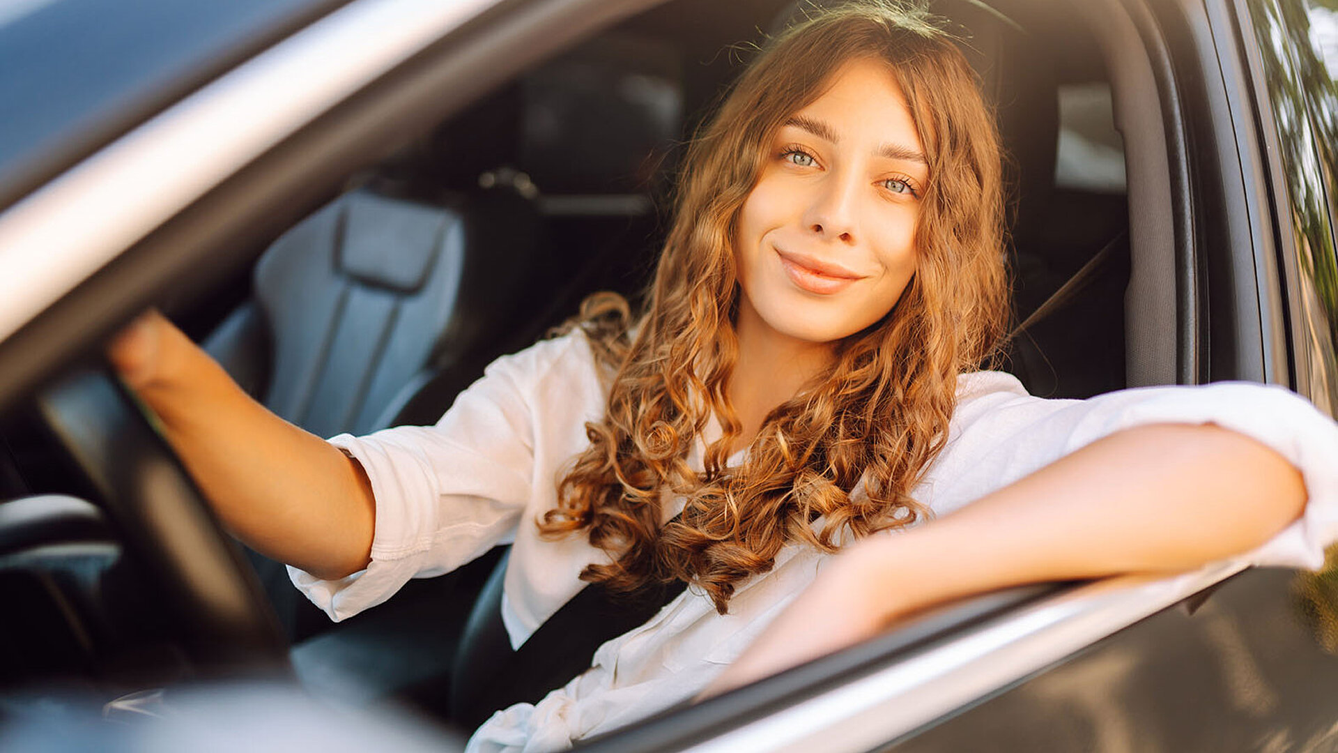 © maxbelchenko-AdobeStock.com Junge Frau sitzt lächelnd am Steuer eines Autos und genießt die Fahrt
