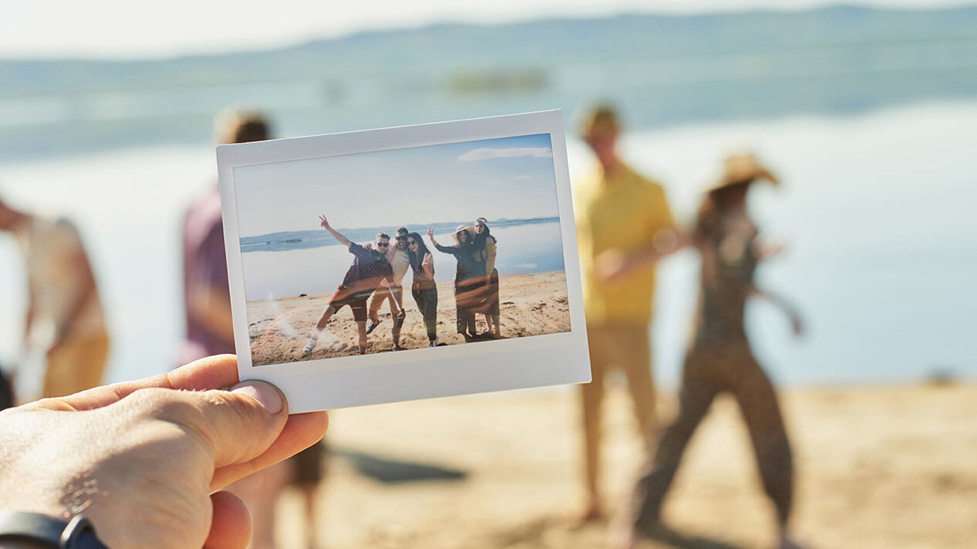 © Pressmaster - AdobeStock.com Hand hält ein Foto am Strand mit Urlaubsszene und Menschen im Hintergrund am Meer