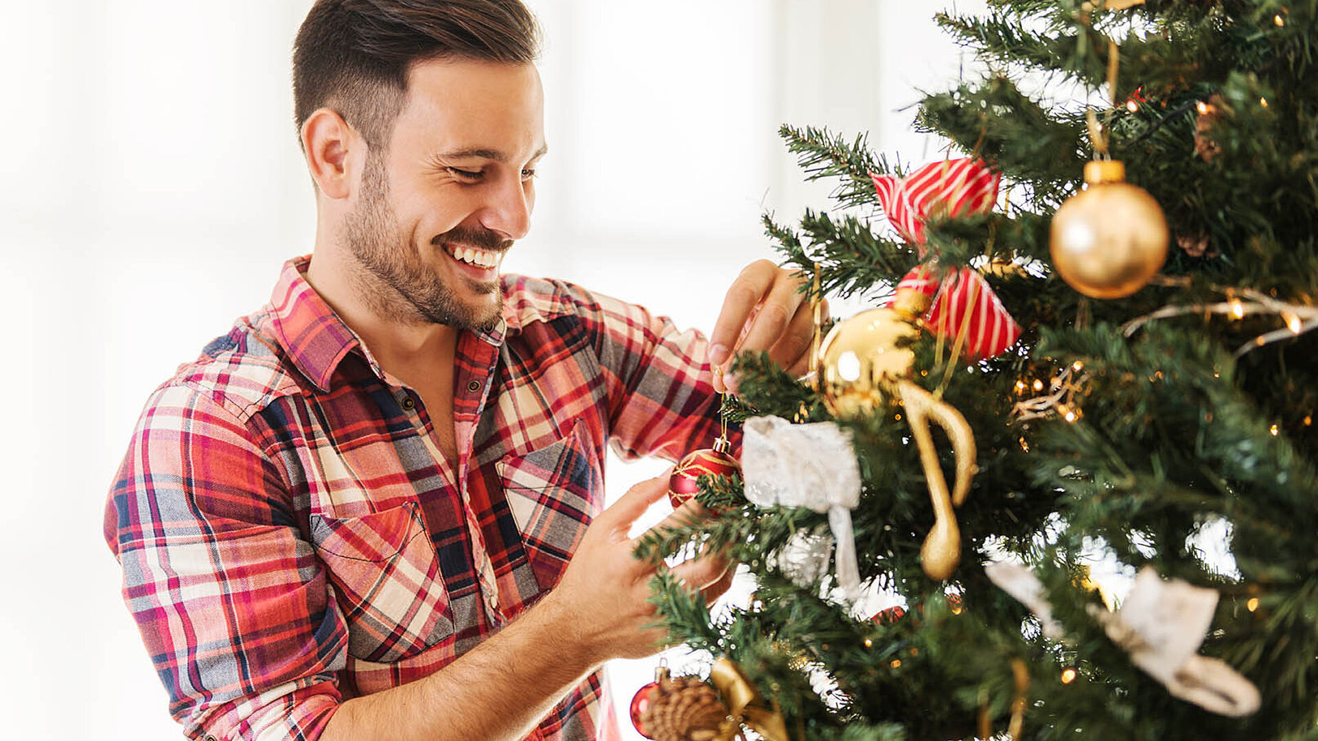 © Tijana - AdobesStock.com Person schmückt einen Weihnachtsbaum mit goldenen und roten Ornamenten und wirkt dabei fröhlich und entspannt.