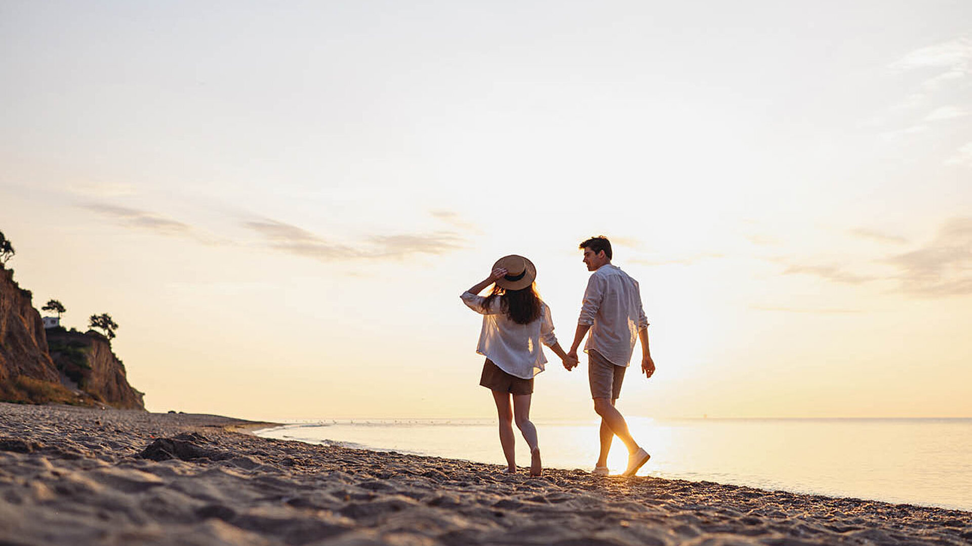 © ViDi_Studio-AdobeStock.com.jpg Paar spaziert Hand in Hand am Strand bei Sonnenuntergang – romantischer Moment am Meer