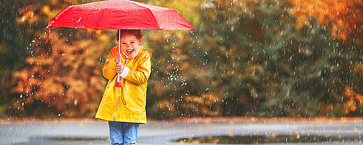 © JenkoAtaman - AdobeStock.com Kind mit gelbem Regenmantel und Stiefeln hält roten Schirm und lacht im herbstlichen Regen.