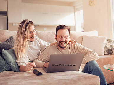 © ivanko80 - AdobeStock.com Paar sitzt entspannt auf dem Sofa und schaut gemeinsam auf Laptop