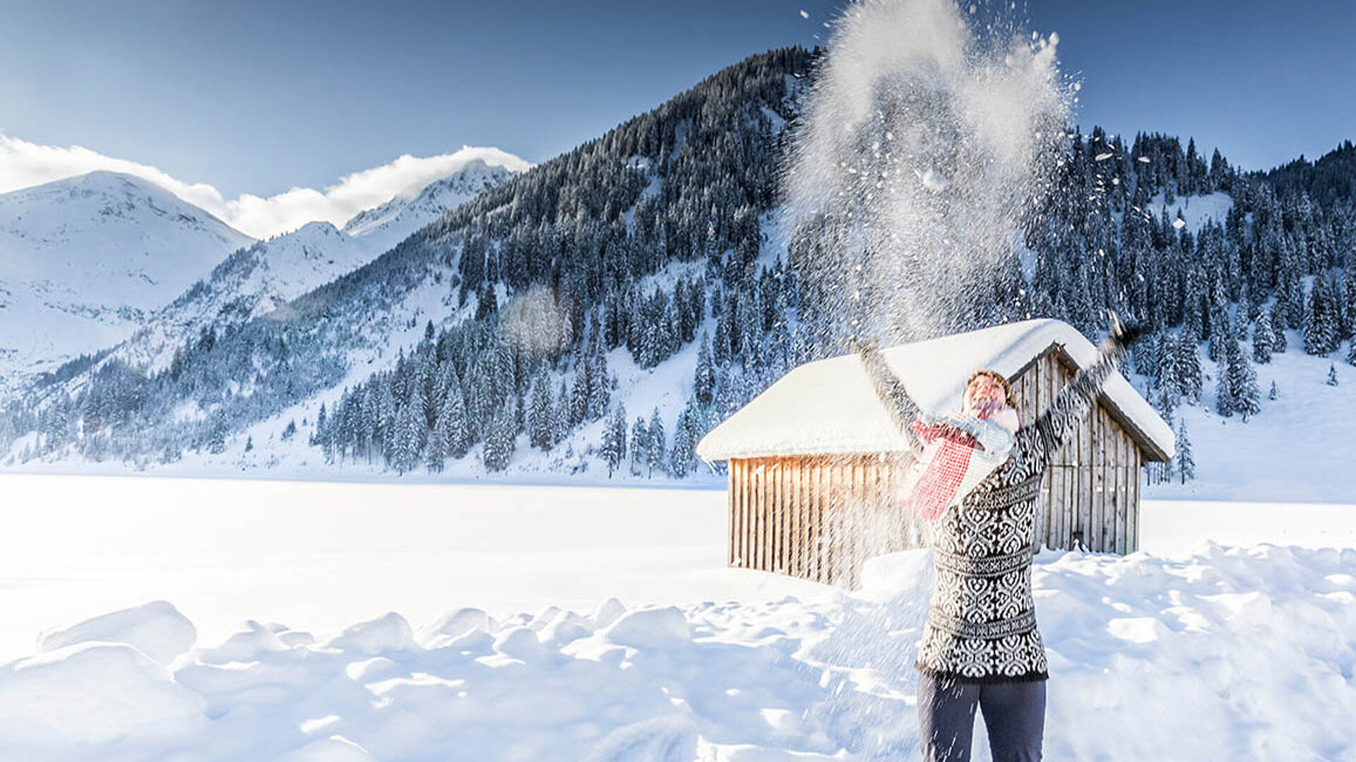 © mmphoto - AdobeStock.com Person wirft Schnee in die Luft vor verschneiten Bergen und einer Hütte in strahlender Winterlandschaft.