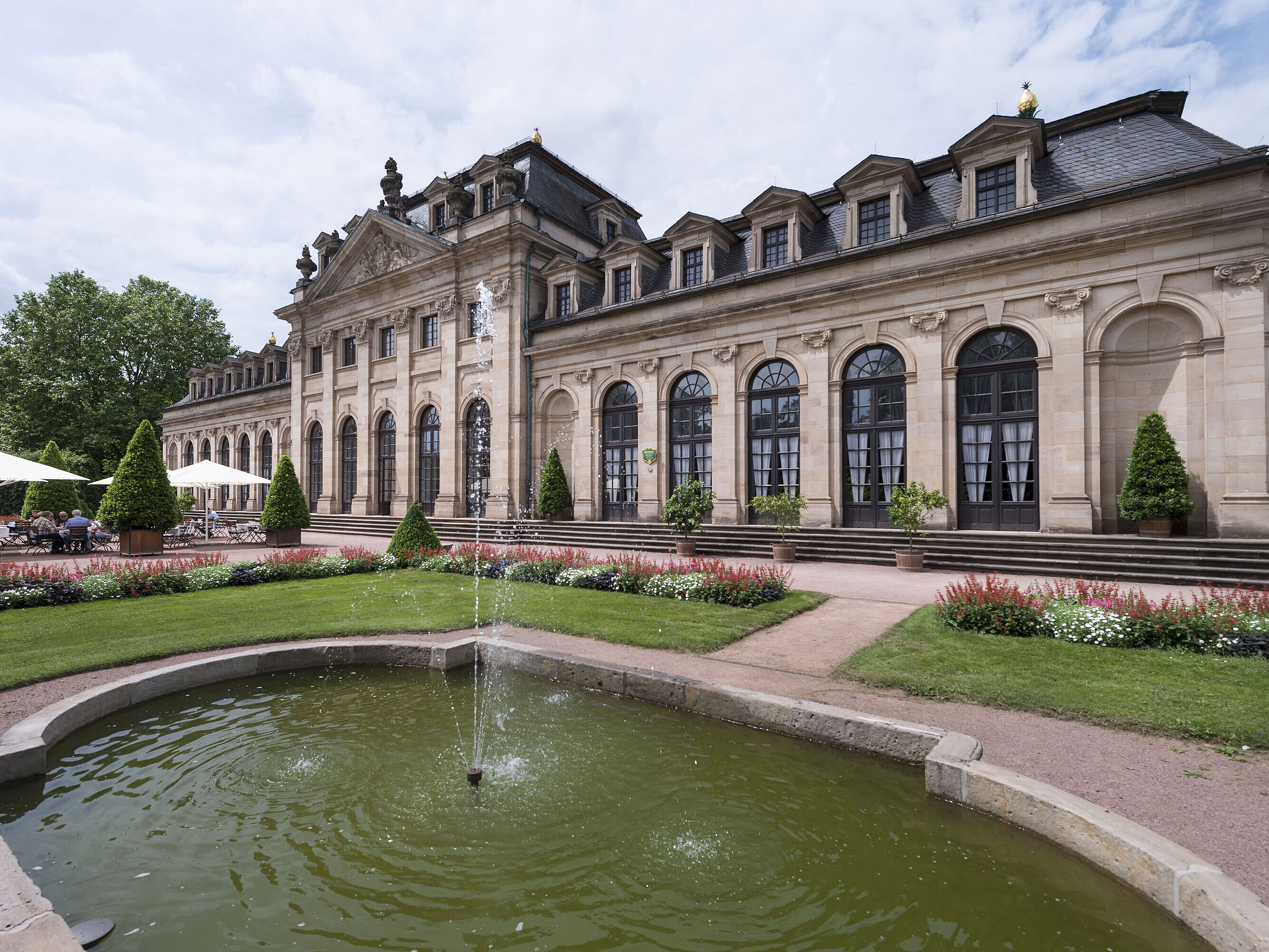 Außenansicht Die barocke Orangerie Fulda mit Terrasse, gepflegtem Garten und historischem Springbrunnen im Vordergrund.