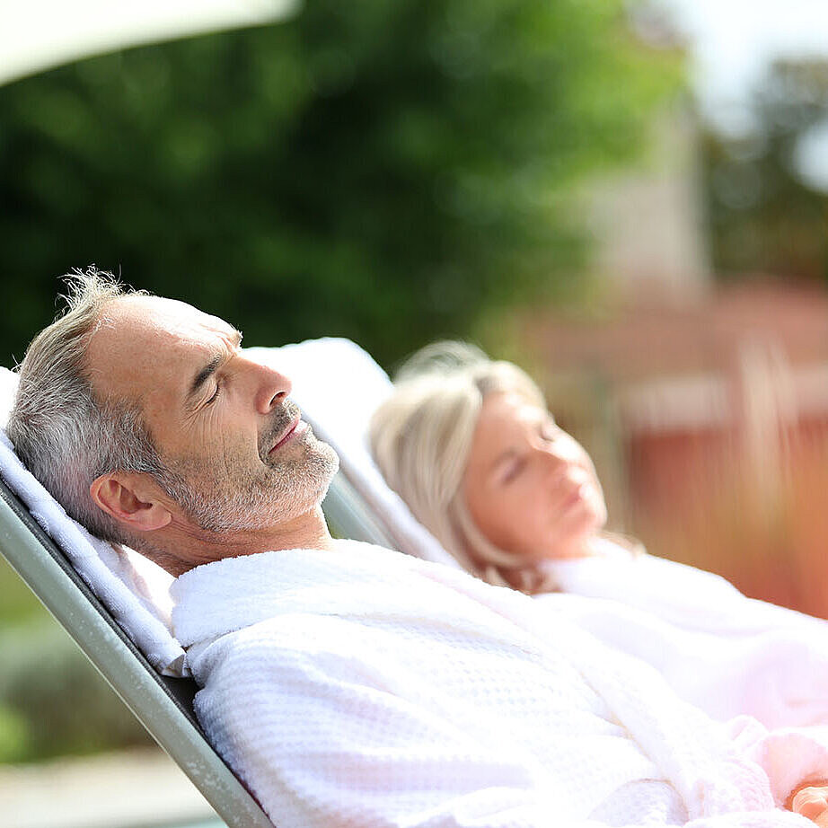 © goodluz - AdobeStock.com Mann und Frau im Bademantel entspannen auf Liegen im Freien und genießen eine erholsame Auszeit.