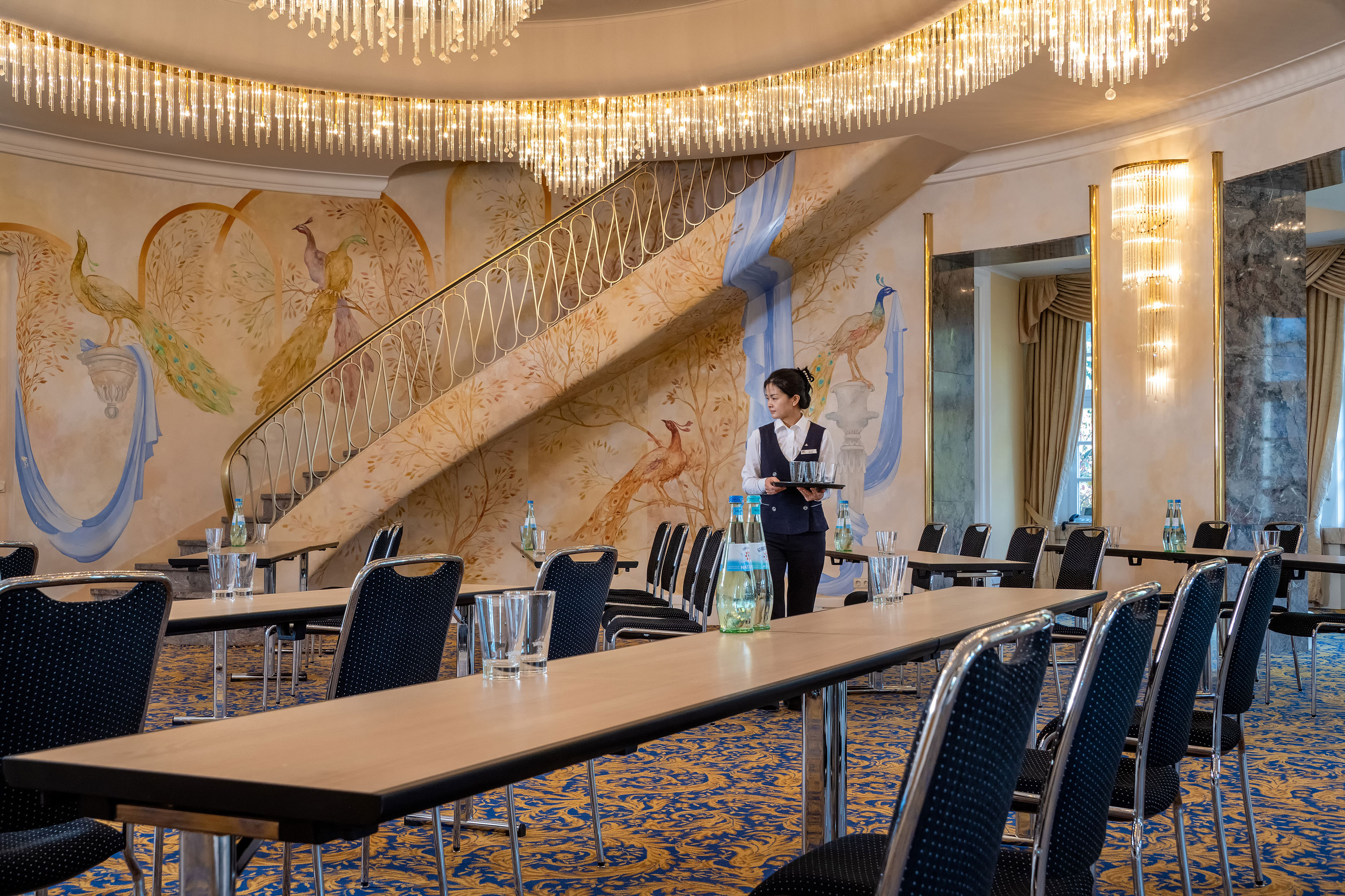 Fürst-von-Pückler-Saal Fürst-von-Pückler-Saal mit Reihenbestuhlung, auffälliger Wandgestaltung, großer Treppe und Servicekraft im Raum.