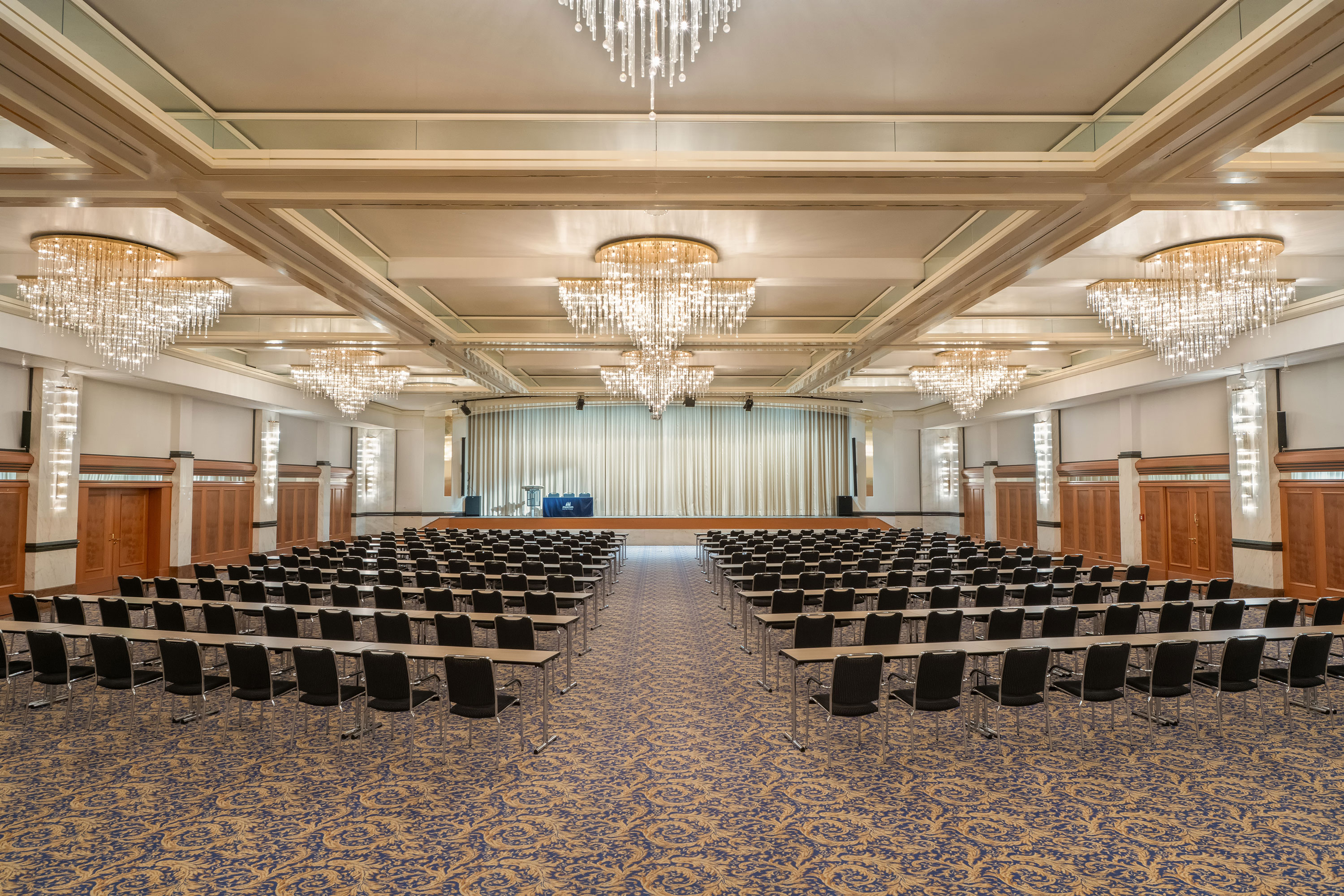 Fürst-von-Metternich-Saal Fürst-von-Metternich-Saal mit großer Bühne, Reihenbestuhlung, eleganten Kronleuchtern und hellem Ambiente.