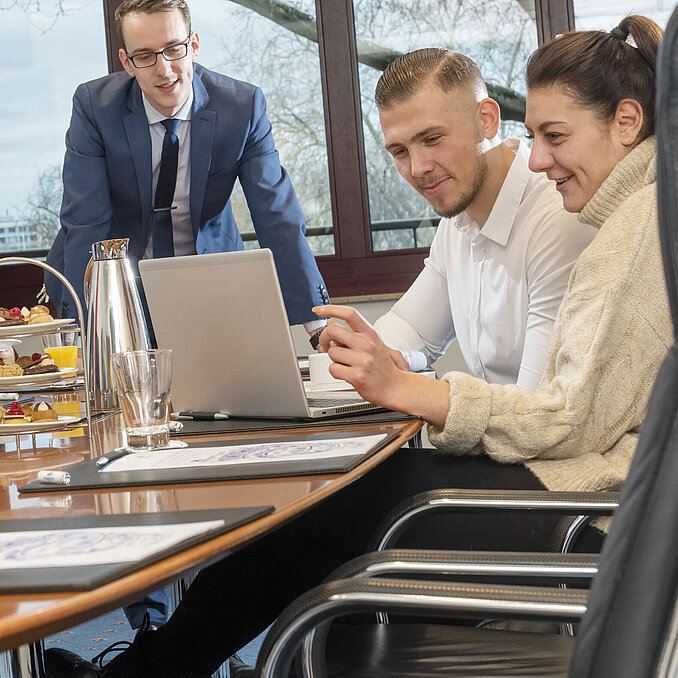 Team in einer Besprechung im Boardroom Maritim Köln, mit Kaffee und Snacks auf dem Tisch, während sie am Laptop arbeiten.