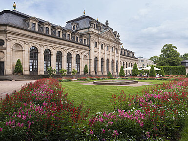 Außenansicht Die historische Orangerie in Fulda mit barocker Fassade, Treppe und blühendem Garten im Vordergrund.